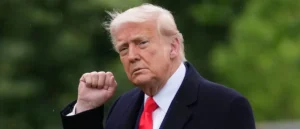 The image shows a person in a suit and red tie, with a fist raised, set against a blurred green background.