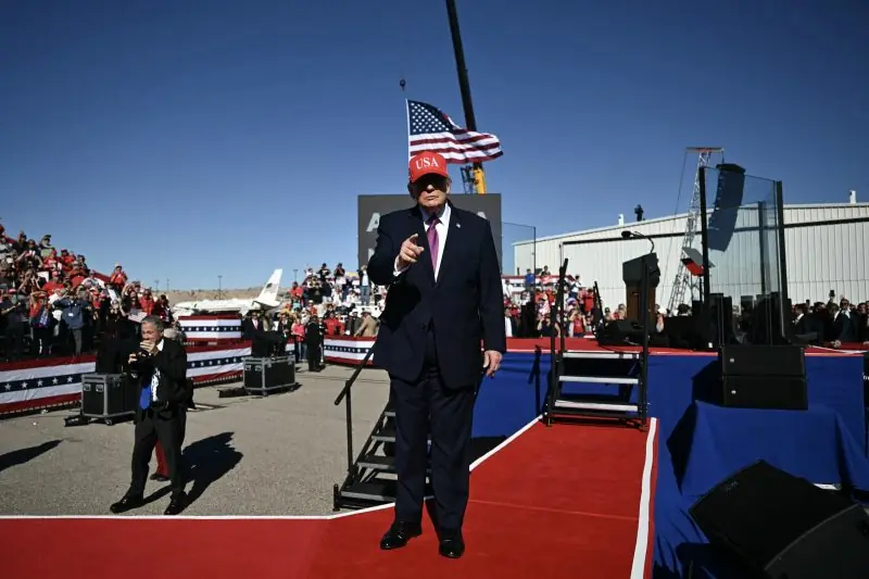 The image shows a political rally with a speaker on stage, wearing a red "USA" hat, addressing a large crowd in a patriotic setting.
