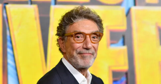 The image shows a man wearing a suit and glasses, standing in front of a colorful backdrop.