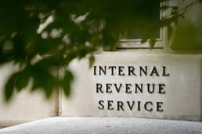 The image shows a sign for the Internal Revenue Service (IRS) partially obscured by green foliage.