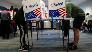 Two individuals are seen bending over at voting booths in a polling station, with "I Voted" signs prominently displayed.