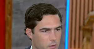 The image shows a man with dark hair, wearing a suit, in what appears to be a news studio setting.