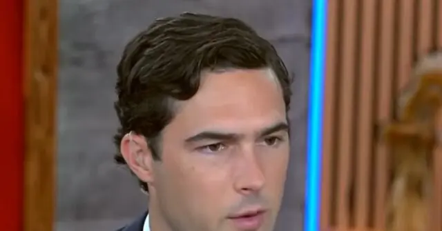 The image shows a man with dark hair, wearing a suit, in what appears to be a news studio setting.