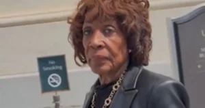 The image shows a person standing in front of a "No Smoking" sign, wearing a dark blazer and a necklace.