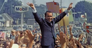 A politician raises his arms in victory as a crowd cheers and waves signs in a celebratory scene.