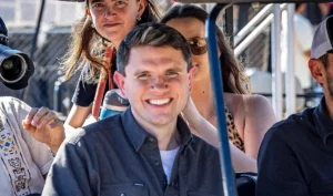 A man smiles while seated among a group of people on a sunny day.