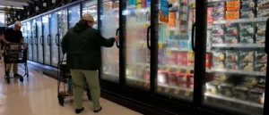 A person is shopping in a grocery store, pushing a cart and reaching for items in a refrigerated section.