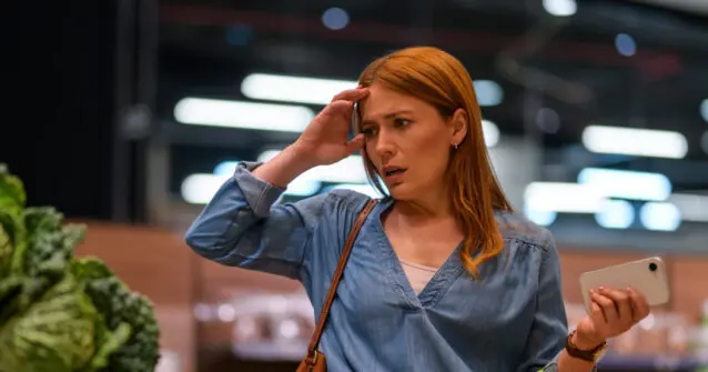 A woman appears to be in a grocery store, holding a phone and looking surprised or confused.