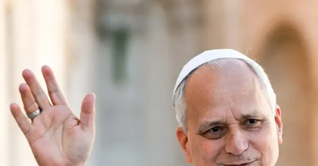 The image shows a person wearing a white head covering, waving with their right hand, in what appears to be an outdoor setting.