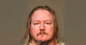 The image appears to be a mugshot of a man with long hair and a mustache, taken against a plain background.