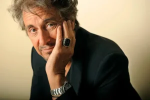 The image shows a person in a dark suit with a hand resting on their face, wearing a ring and a wristwatch.