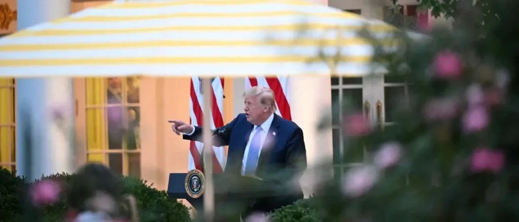 The image shows a person speaking at a podium with an American flag in the background, under a striped canopy.