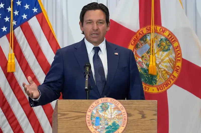 A man in a suit stands at a podium with the Great Seal of the State of Florida and an American flag in the background, delivering a speech.