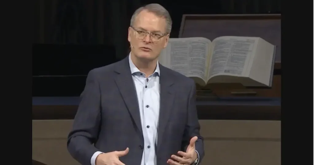 The image shows a person in a suit gesturing while speaking, with an open book in the background, suggesting a formal or educational setting.