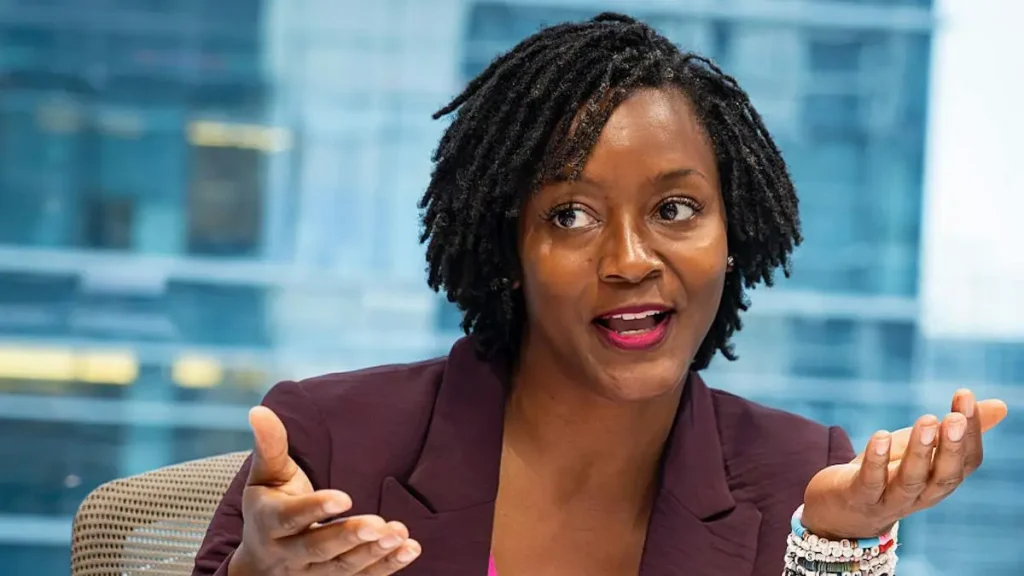 A woman in a professional setting gestures with her hands while speaking, suggesting she is engaged in a discussion or presentation.
