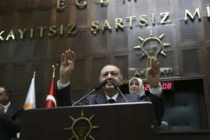 The image shows a person standing at a podium with their hands raised, in front of a backdrop featuring the text "KAYITSIZ ŞARTSIZ Mİ" and a logo, with flags and a clock visible in the background.