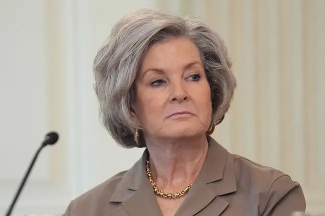 The image shows a woman with gray hair wearing a brown suit and a gold necklace, seated in front of a microphone, likely during a formal or professional setting.