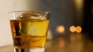 A glass of amber-colored liquid with ice cubes sits on a wooden surface, with blurred lights in the background.