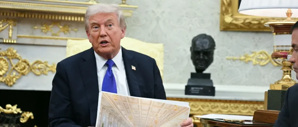 The image shows a man in a suit holding a document and speaking in a room with ornate decor, including a bust and gold accents.