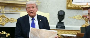The image shows a man in a suit holding a document and speaking in a room with ornate decor, including a bust and gold accents.