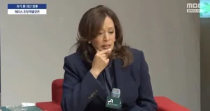 The image shows a person in a dark suit sitting on a red chair, speaking into a microphone, with text in Korean and English indicating a news segment.