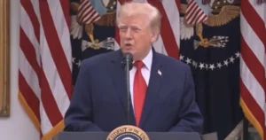 The image shows a person speaking at a podium with the seal of the President of the United States and American flags in the background.