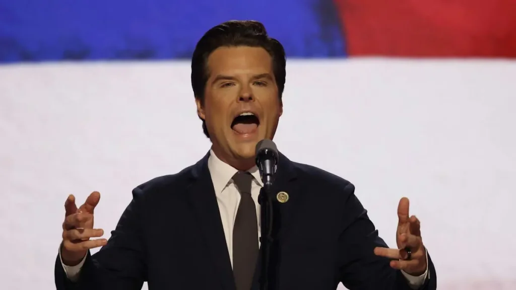 A man in a suit and tie is speaking passionately into a microphone, with an American flag in the background.