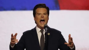 A man in a suit and tie is speaking passionately into a microphone, with an American flag in the background.