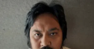 The image shows a close-up of a person with dark hair and a mustache, set against a plain background.