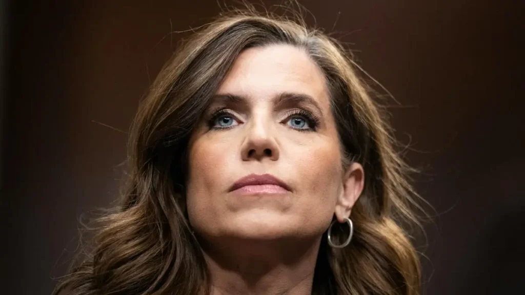 The image shows a person with long, wavy hair and a serious expression, wearing a large hoop earring.