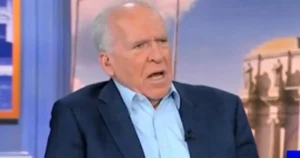 The image shows a man in a suit speaking on a news program, with a backdrop featuring a building and a blue sky.