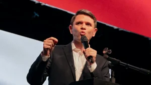 A man in a suit is speaking into a microphone at a podium, gesturing with his hand.