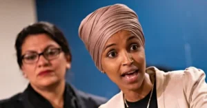 The image shows two women speaking at a press conference, with one wearing a beige headscarf and the other in a black outfit.