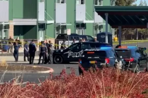 Police officers are gathered around a crime scene at a building with a police car and other vehicles present.