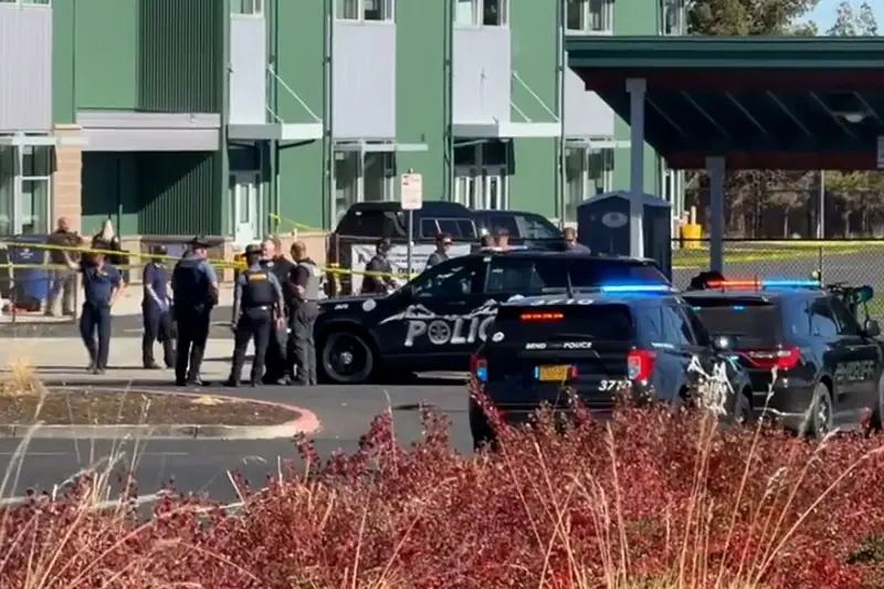 Police officers are gathered around a crime scene at a building with a police car and other vehicles present.
