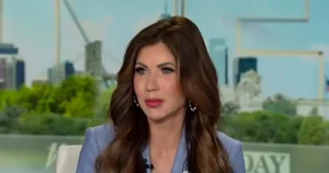 The image shows a news anchor in a studio setting with a cityscape background.