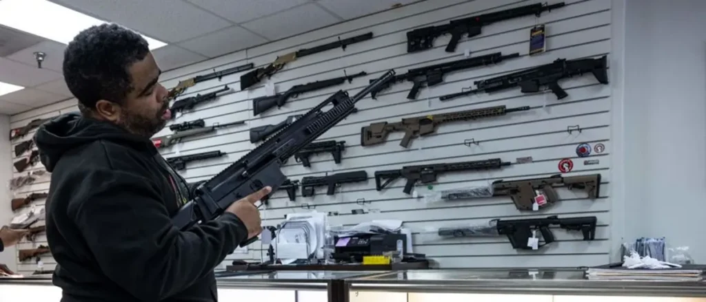 A man examines a firearm in a store displaying various rifles on a wall.