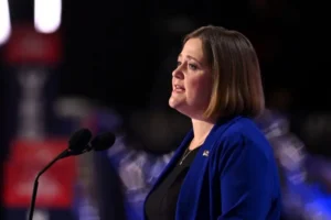 A woman in a blue blazer is speaking at a podium in front of a blurred audience.