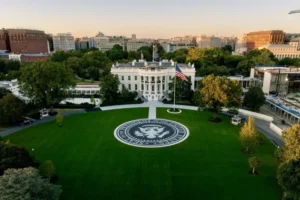 The image shows an aerial view of the White House with the presidential seal prominently displayed on the lawn in front of it.