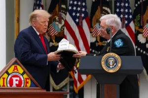 The image shows a formal event where a person is presenting a firefighter's helmet to another individual at a podium with the seal of the President of the United States in the background.