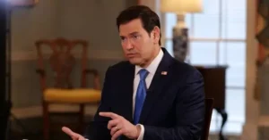 The image shows a man in a suit and tie, seated and gesturing with his hands, likely during an interview or news segment.