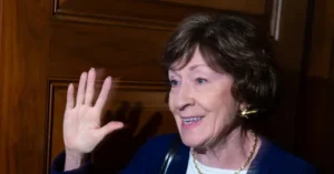 The image shows a person waving with a smile, wearing a blue jacket and a gold necklace, in front of a wooden door.