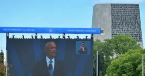The image captures the grand opening ceremony of the Obama Presidential Center, with a large screen displaying a speaker and a smaller inset showing another individual.