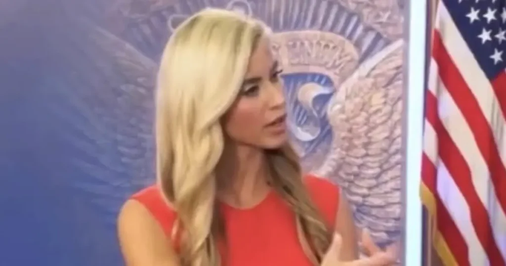 The image shows a person in a red dress speaking in front of an American flag and the U.S. presidential seal.