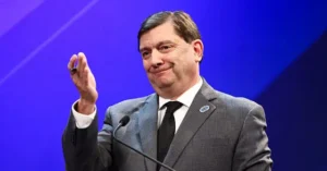 The image shows a man in a suit gesturing with his right hand while speaking at a podium, set against a blue background.