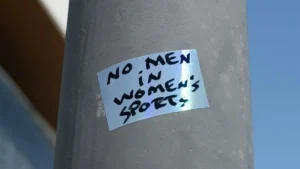 The image shows a sticker on a pole that reads "NO MEN IN WOMEN'S SPORTS."