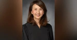 The image shows a person wearing a black robe, likely a judge or academic, against a neutral background.