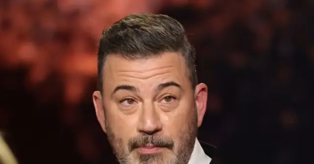 The image shows a close-up of a man with short, styled hair and a beard, wearing a suit, against a blurred background.