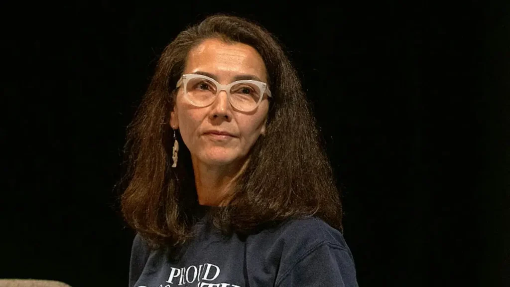 The image shows a person wearing a dark blue sweatshirt with the text "PROUD" visible, sitting against a dark background.