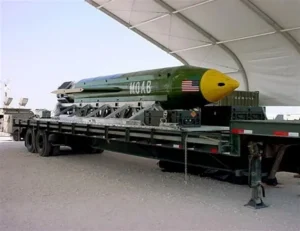 The image shows a large military bomb, labeled "MOAB," being transported on a flatbed truck under a tent.